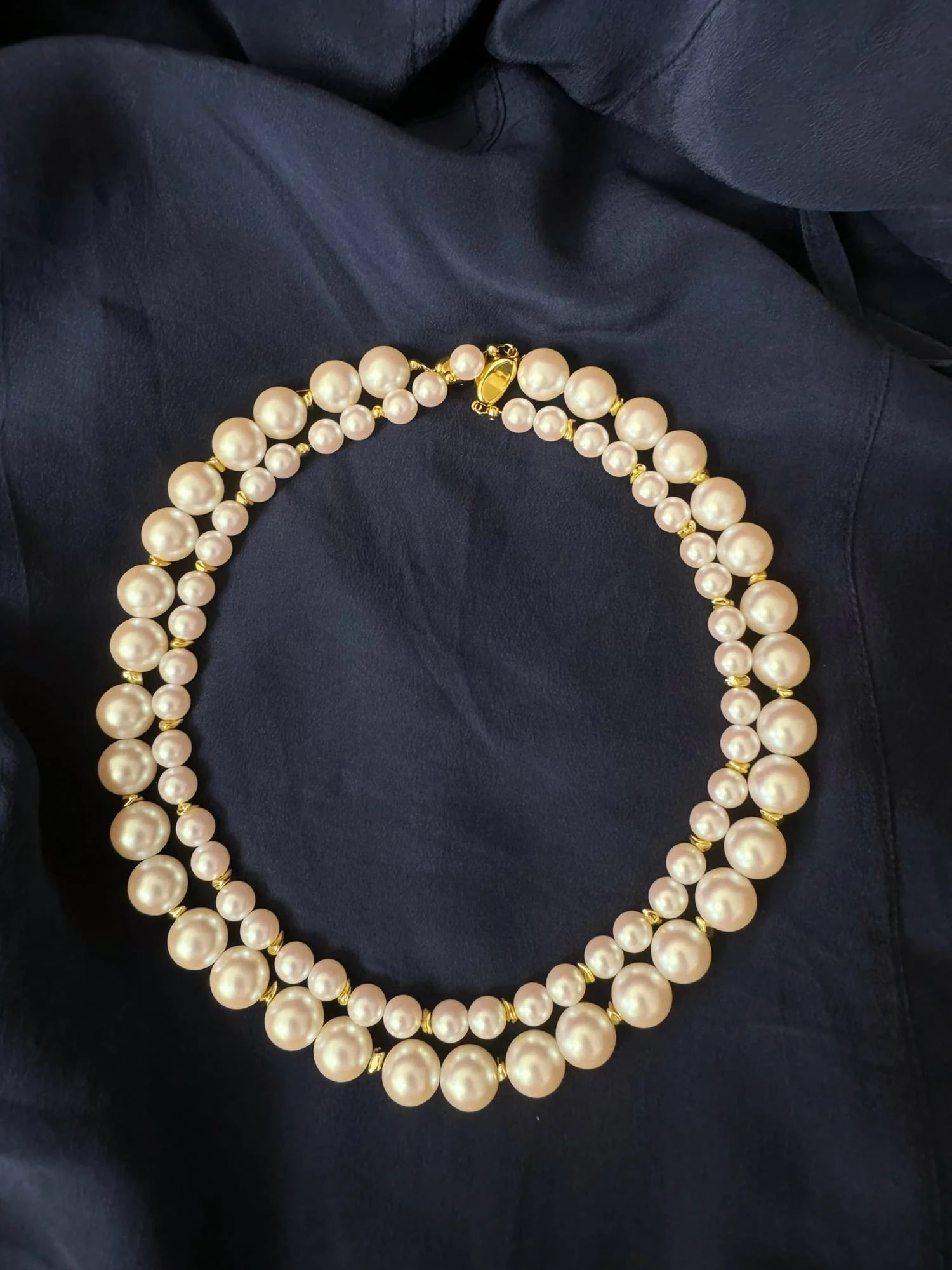 The double-strand white-bead necklace laid in a circle on navy fabric, outer beads graduating larger toward the centre, gold-toned oval box clasp at the top.