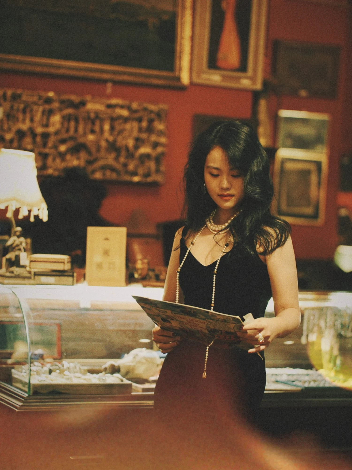 The brown bead long necklace on a model looking through a folio in a red-walled antique shop, its drop visible at the end.