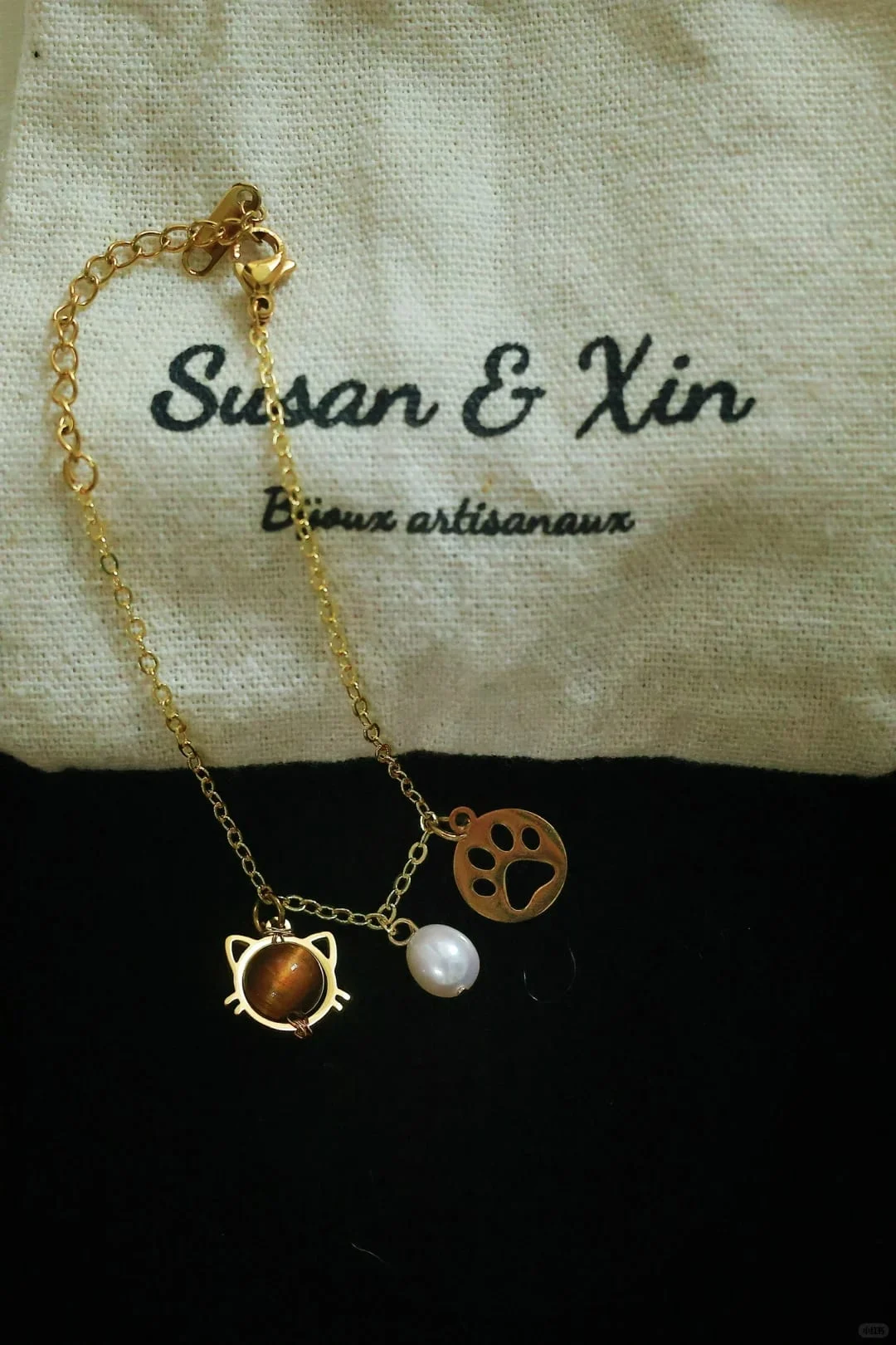 Fine gold-toned chain bracelet with a cat-face frame holding a brown bead, a white iridescent oval bead and an openwork paw-print disc, with lobster clasp and extender, on a Susan&Xin pouch against black.