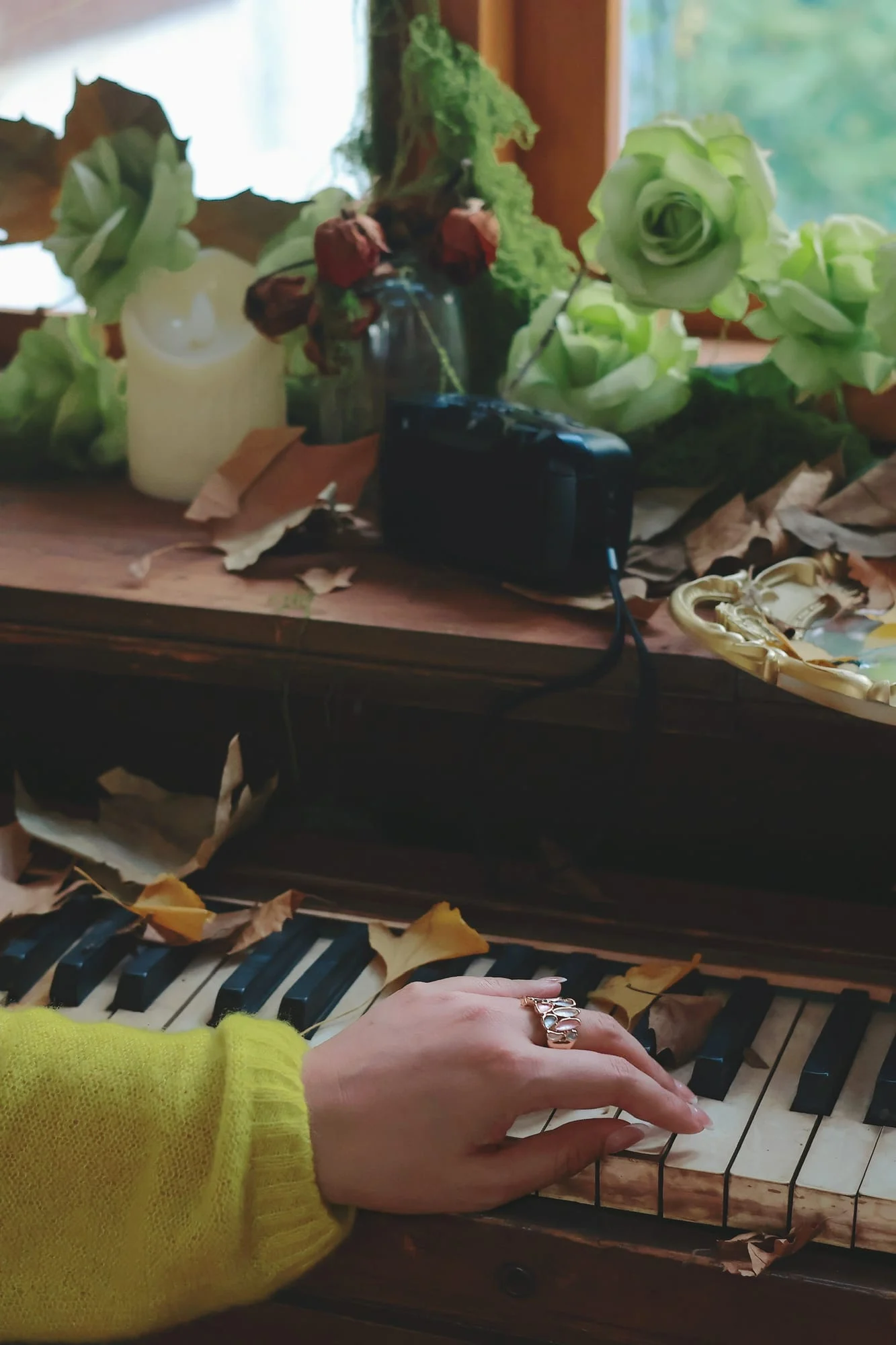 Hand wearing the wide gold-toned enamel leaf ring on the keys of an old piano scattered with autumn leaves, green roses and a candle by the window, yellow knit sleeve.