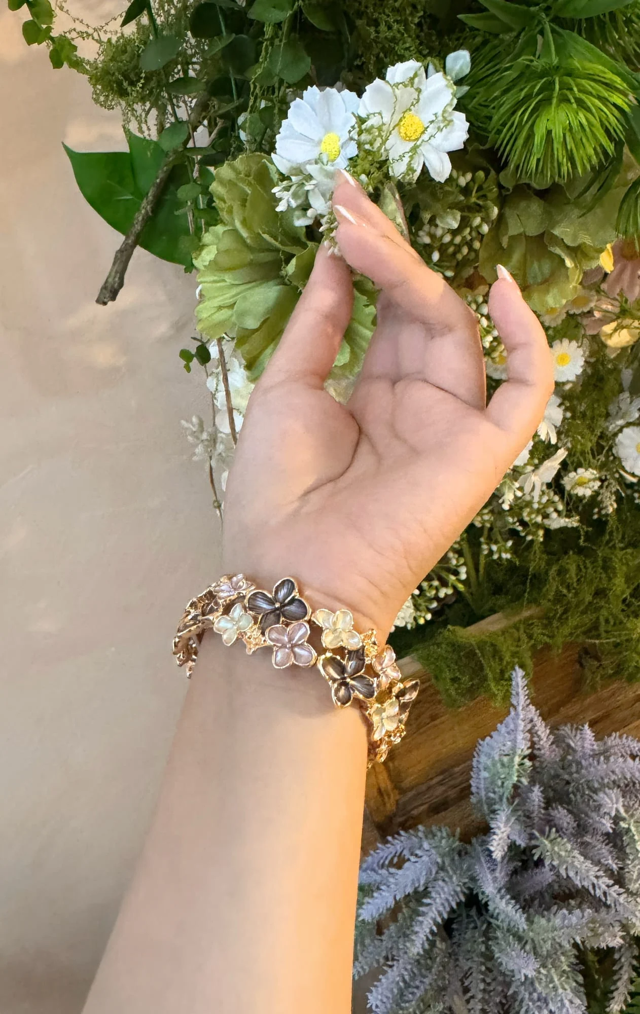 Wrist wearing the gold-toned four-petal enamel flower bracelet, fingers touching white daisies and greenery.