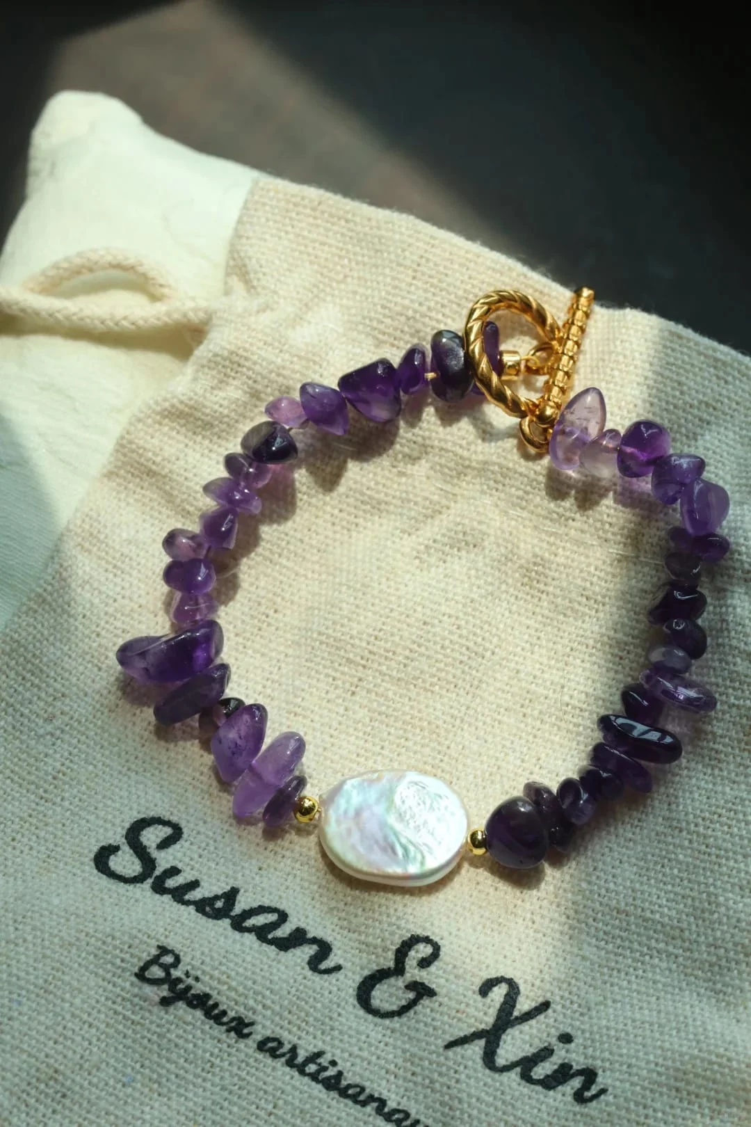 Bracelet of irregular violet beads with a flat white iridescent centrepiece and a gold-toned toggle clasp, resting on a Susan&Xin fabric pouch.