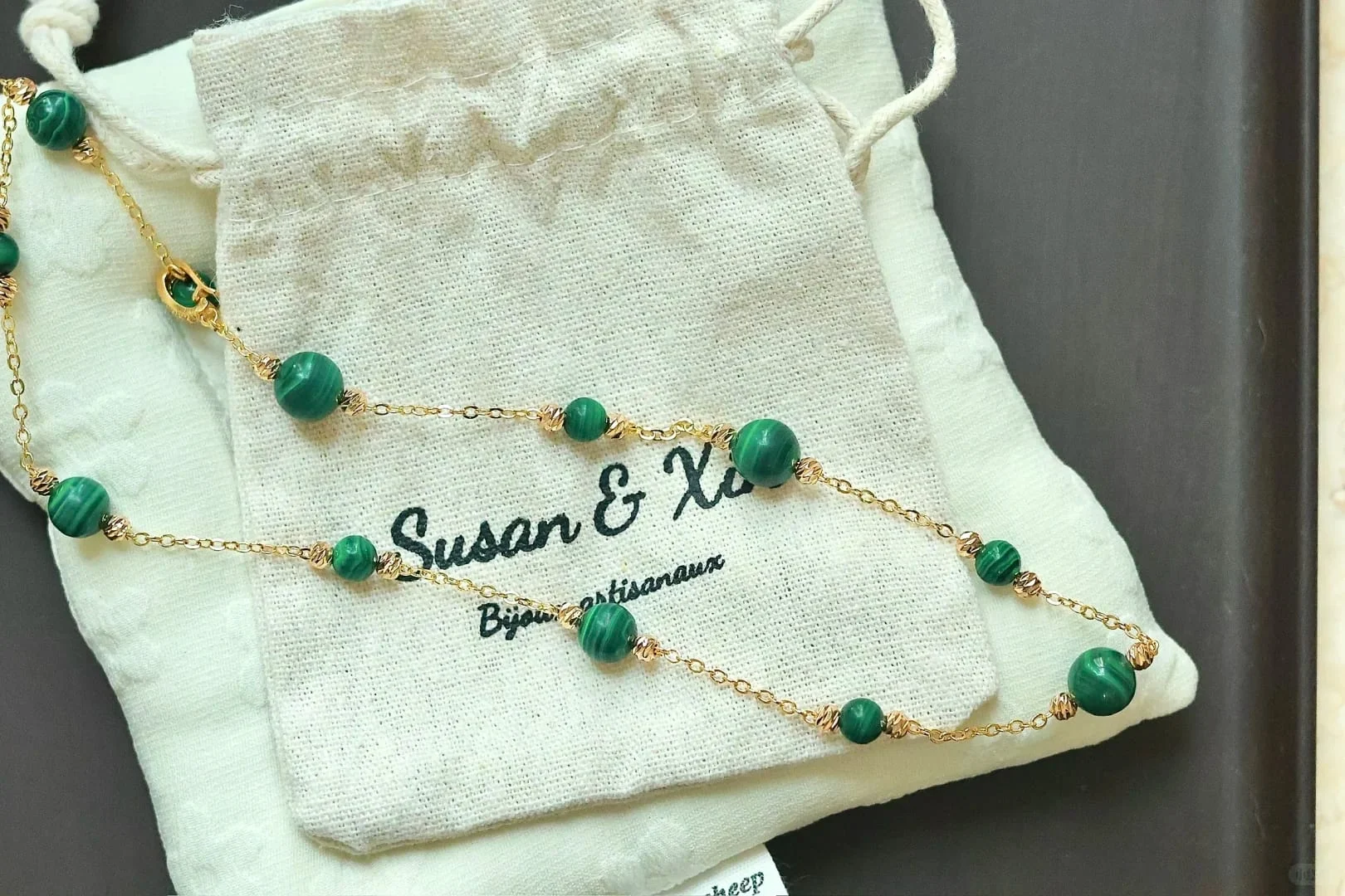 Fine gold-toned chain necklace with spaced banded malachite beads, each between small faceted gold-toned beads, looped over a Susan&Xin fabric pouch.