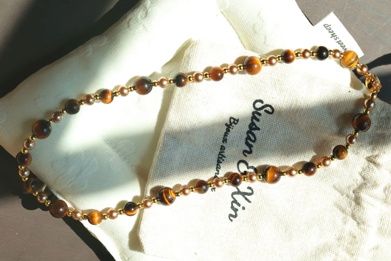 Necklace of round tiger’s eye beads alternating with smaller bronze-toned beads and gold-toned spacers, with a gold-toned clasp, resting on a Susan&Xin fabric pouch.