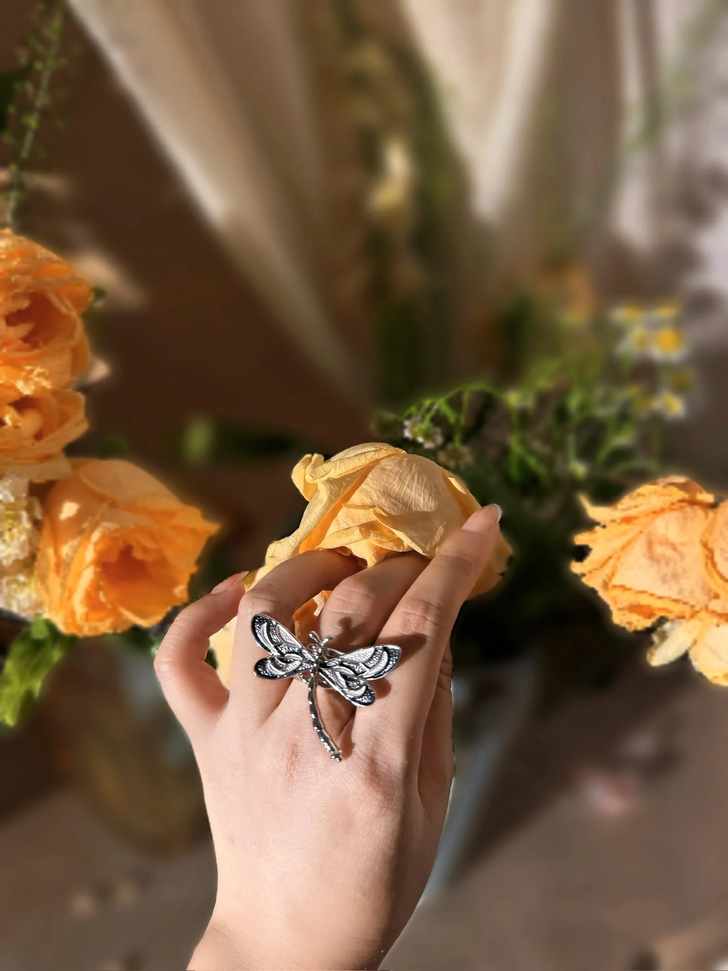 Hand wearing the silver-toned dragonfly ring with black and white enamel wings, fingertips on a dried apricot rose, bouquet behind.