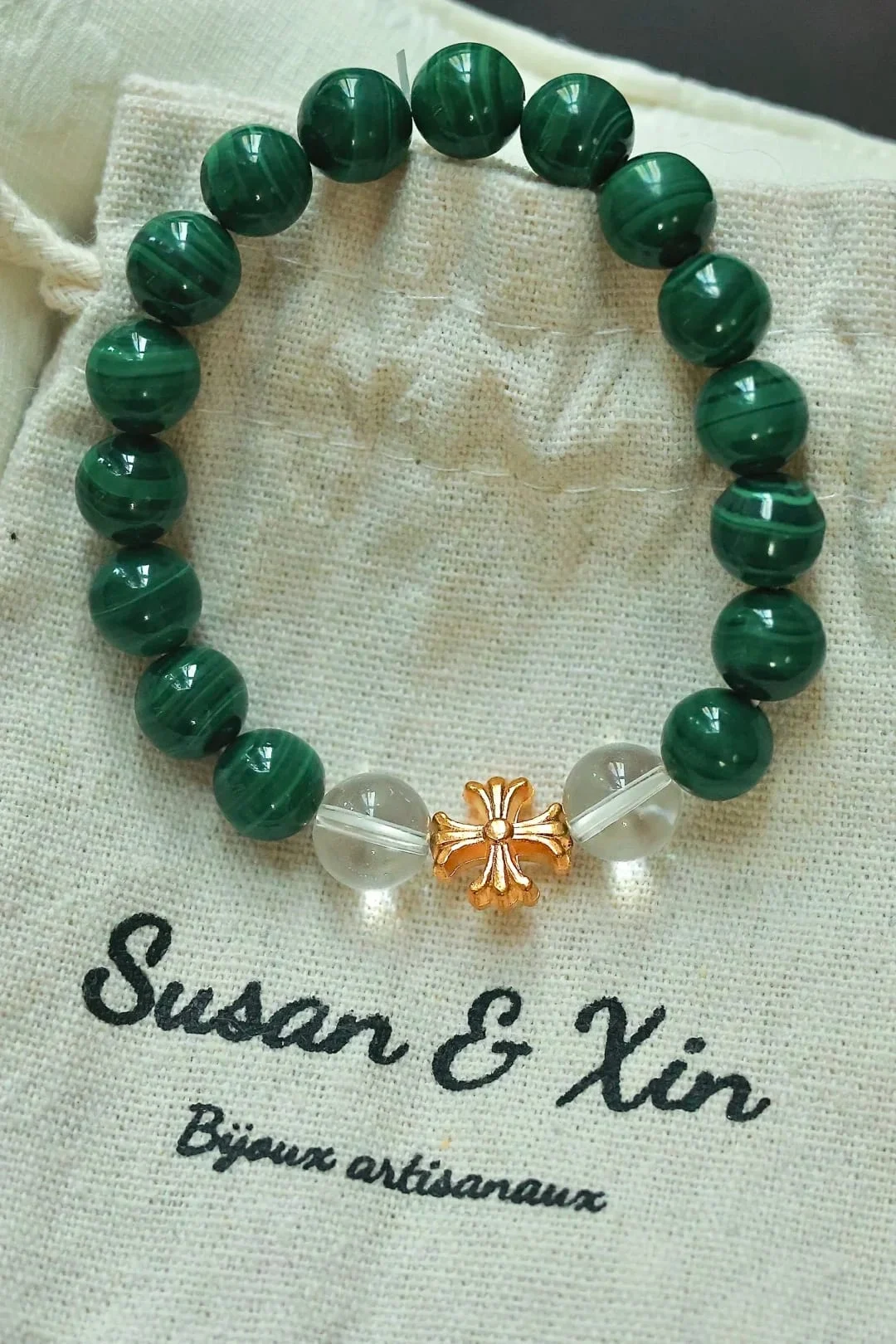 Bracelet of banded malachite beads with a gold-toned cross-shaped bead between two clear round beads, on a Susan&Xin fabric pouch.