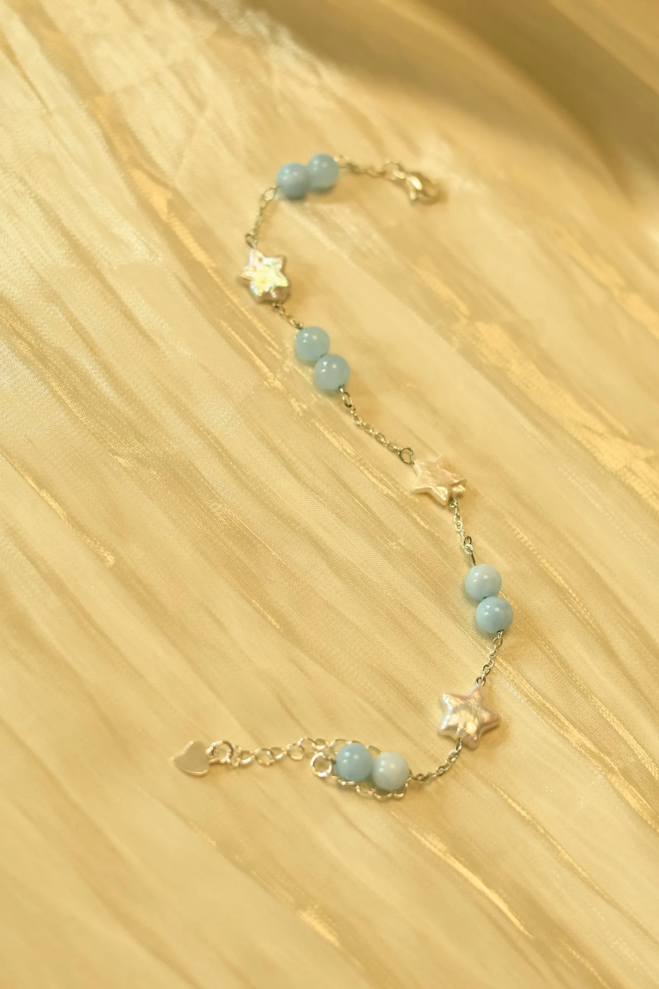 Fine silver-toned chain bracelet with pairs of pale-blue beads and white iridescent stars, lobster clasp, extender chain and heart tag, on gold satin.