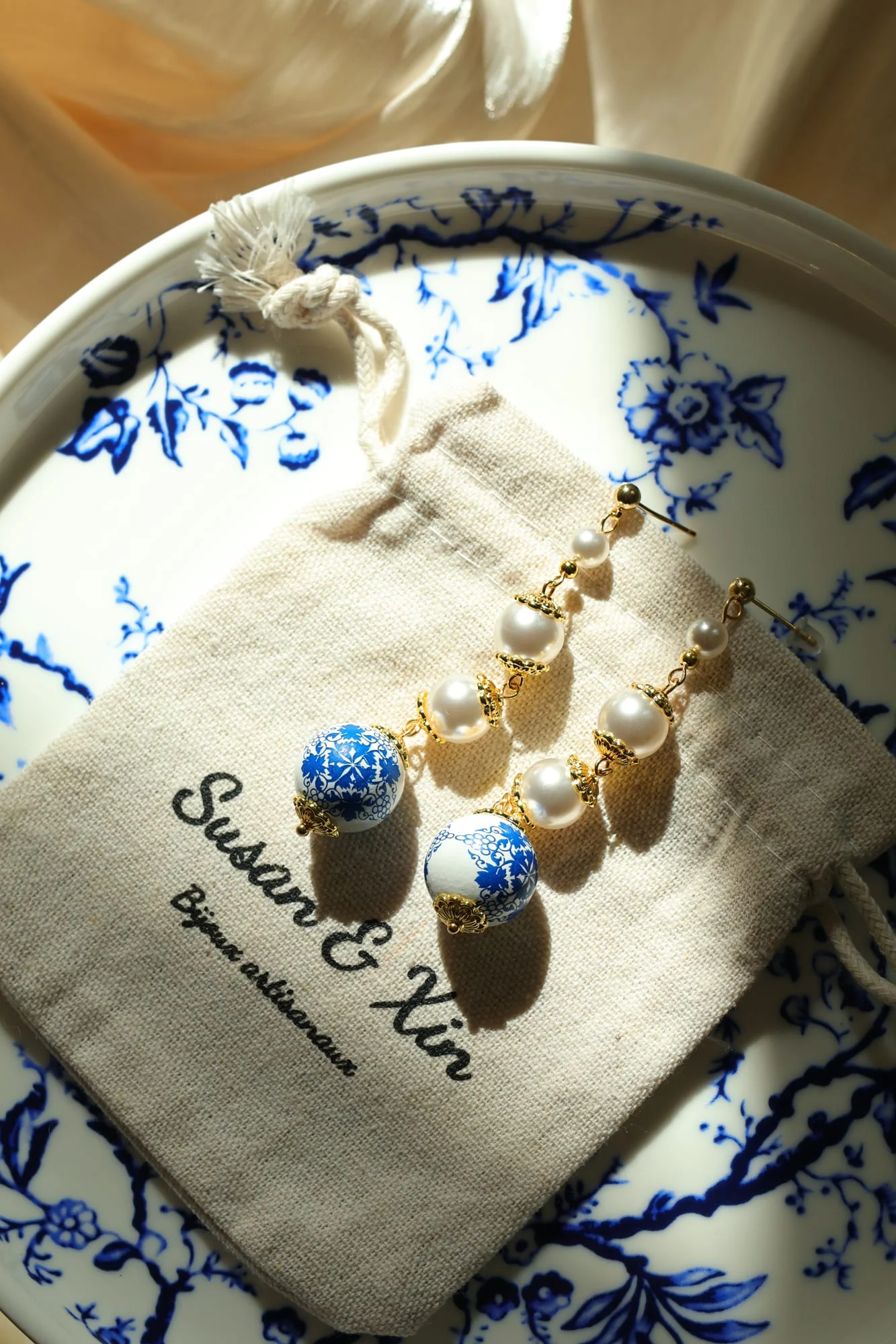 Pair of long drop bead earrings on a Susan&Xin pouch on a blue floral plate in slanting sunlight: white beads in a row ending in blue-and-white patterned beads.