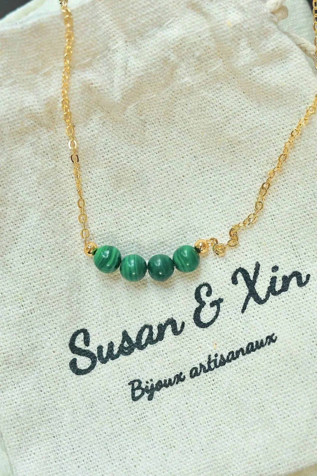 Fine gold-toned chain necklace with four banded malachite beads at the centre and a small gold-toned bead at each end, on a Susan&Xin fabric pouch.