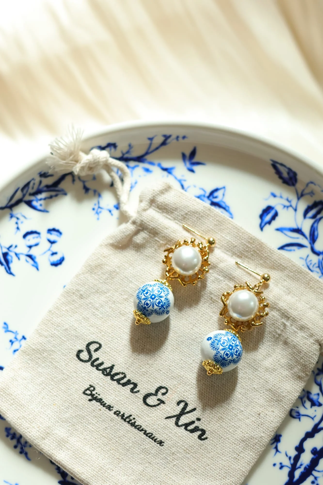 Pair of framed bead drop earrings on a Susan&Xin pouch set on a blue floral plate: white beads in gold-toned openwork frames with blue-and-white patterned beads below.