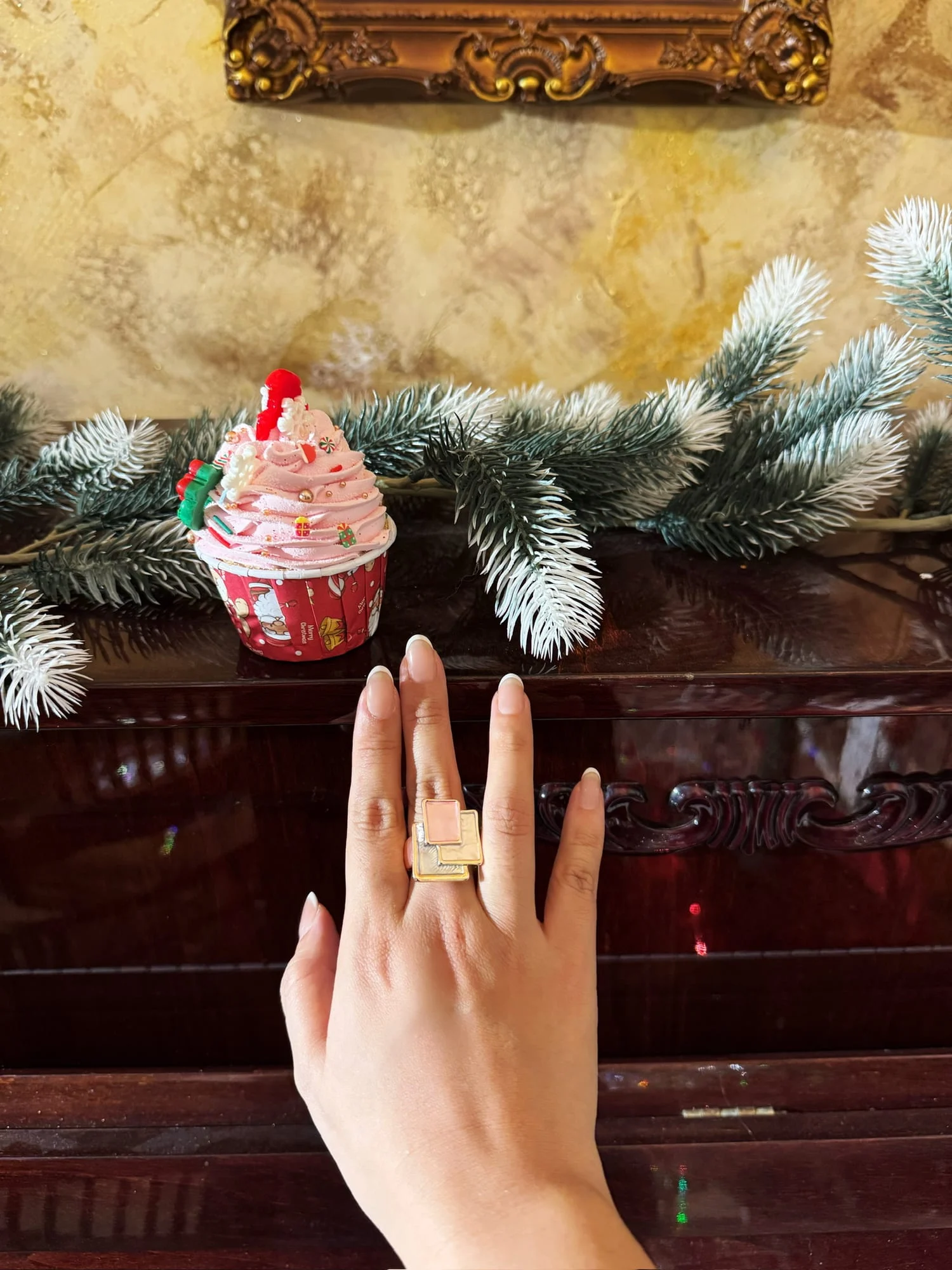 Hand wearing the gold-toned stacked square ring with a pink enamel tile, resting on a dark piano lid with a Christmas cupcake ornament and fir branches.