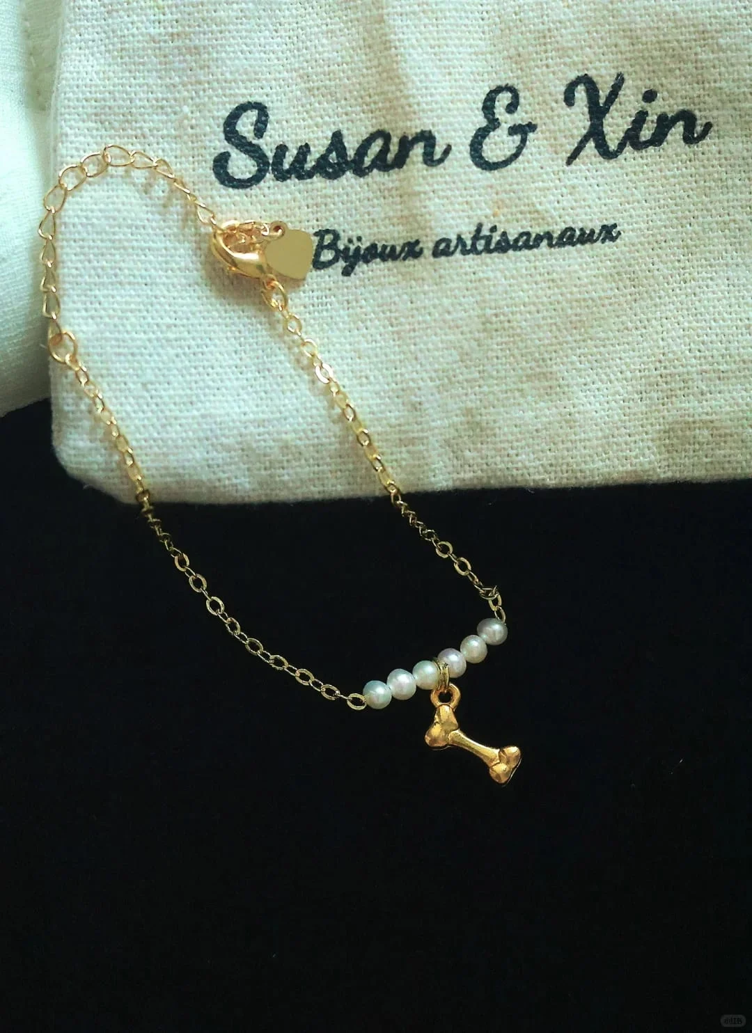 Fine gold-toned chain bracelet with four small white beads and a gold-toned bone charm, lobster clasp, extender chain and heart tag, on a Susan&Xin pouch against black.