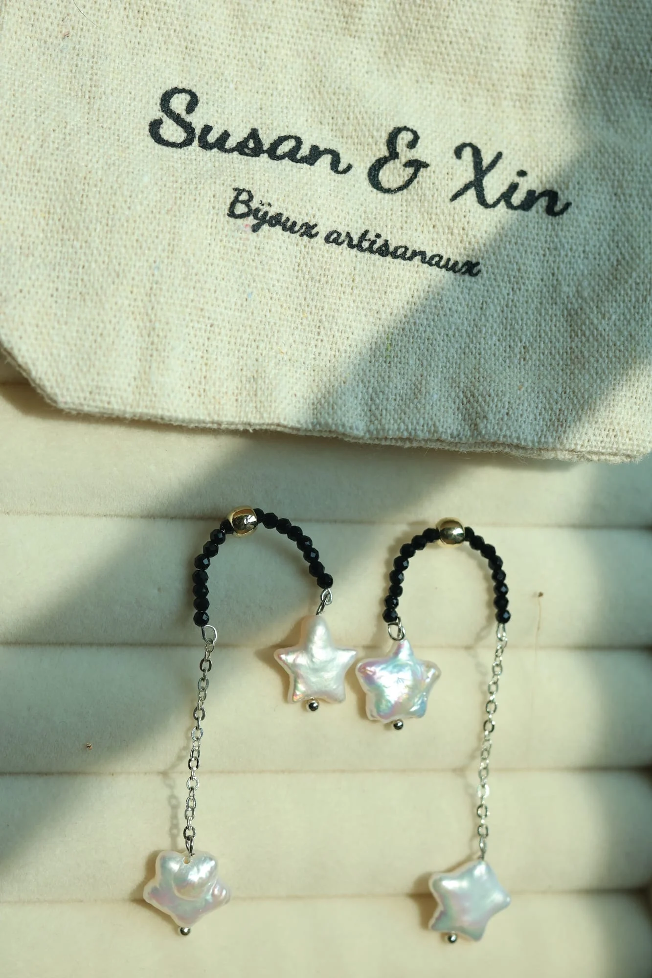 Pair of earrings laid on a jewellery tray: arcs of tiny faceted black beads with a gold-toned bead on top, each dropping two white iridescent stars at different lengths; a Susan&Xin pouch above.