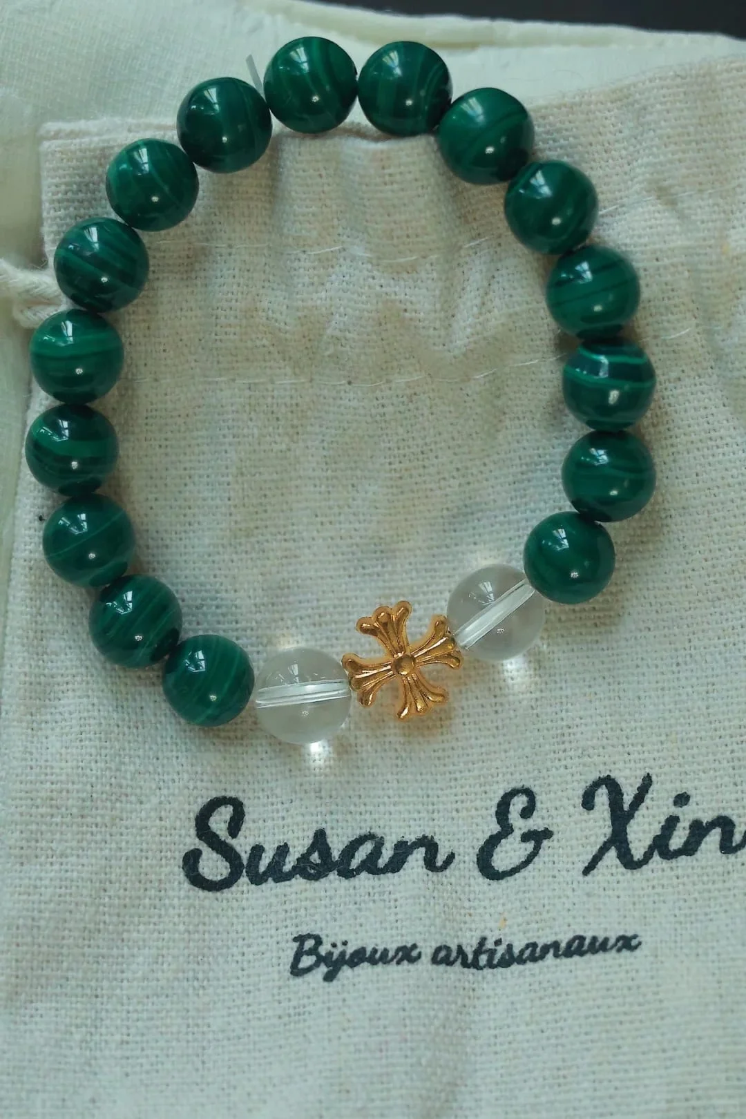 Another photograph of the same malachite cross bead bracelet, gold-toned cross and two clear beads at the bottom, on a Susan&Xin pouch.