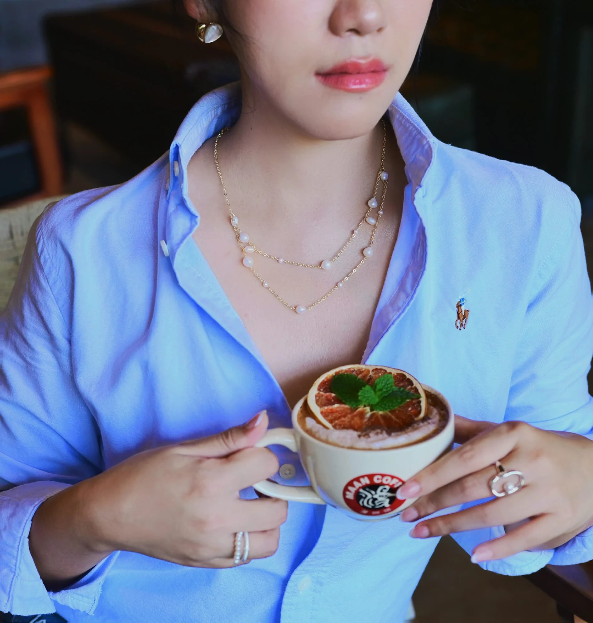 Two white-bead station necklaces layered on a model in a pale-blue shirt holding a coffee cup; this is the upper, larger-beaded one.