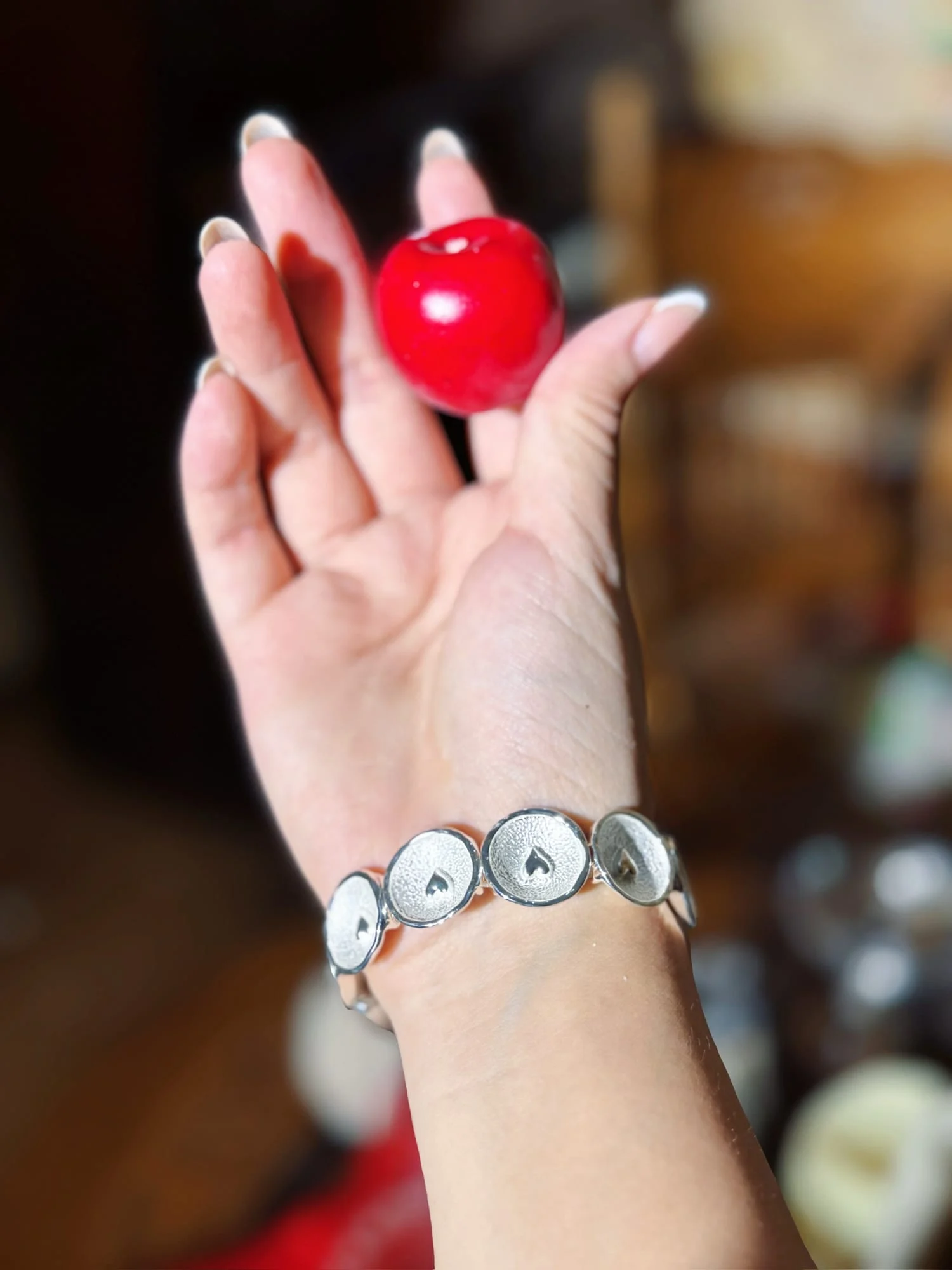 Wrist wearing the silver-toned heart disc bracelet, hand holding a red cherry ornament, background blurred.