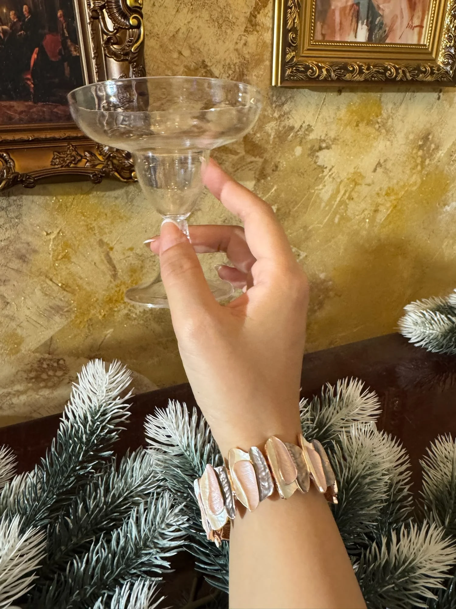 Wrist wearing the wide petal plate bracelet in pink, silver-grey and gold tones, hand raising a clear coupe glass, gilded wall, picture frames and fir branches behind.