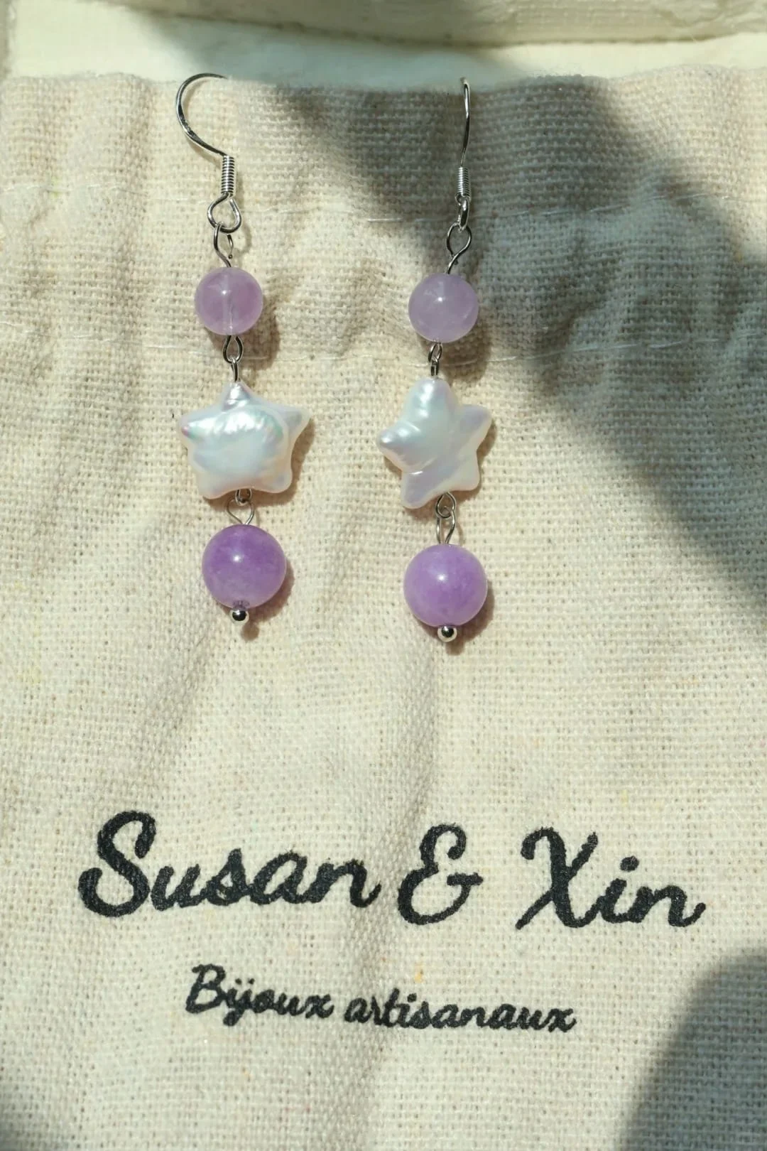 Pair of silver-toned hook earrings with white iridescent stars between lilac and violet round beads, on a Susan&Xin fabric pouch.