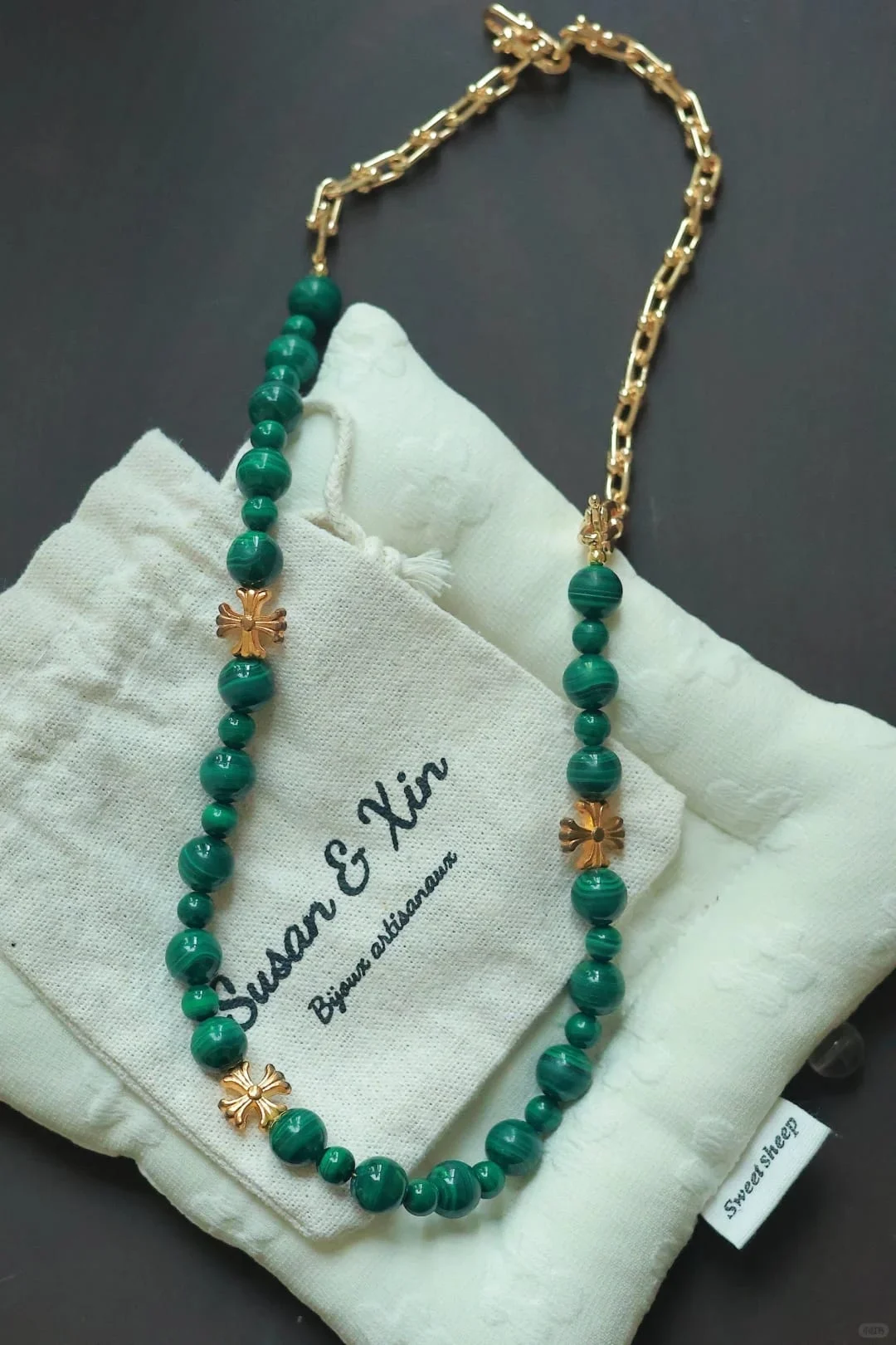 The same malachite cross and chain necklace laid on a Susan&Xin pouch, chunky U-link chain at the top, dark background.