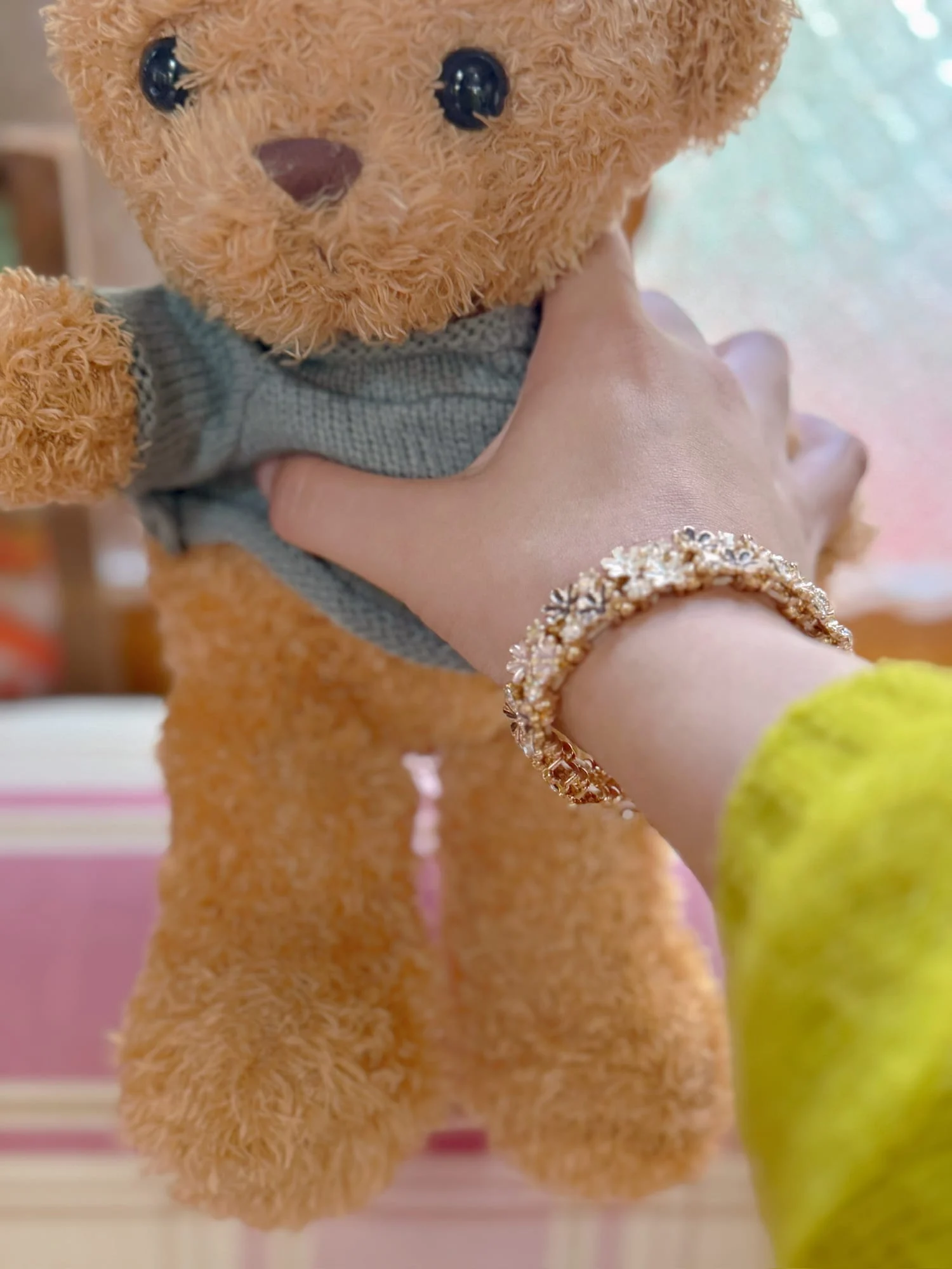 Wrist wearing the gold-toned small enamel flower bracelet, hand holding a teddy bear in a grey sweater, yellow-green knit sleeve.