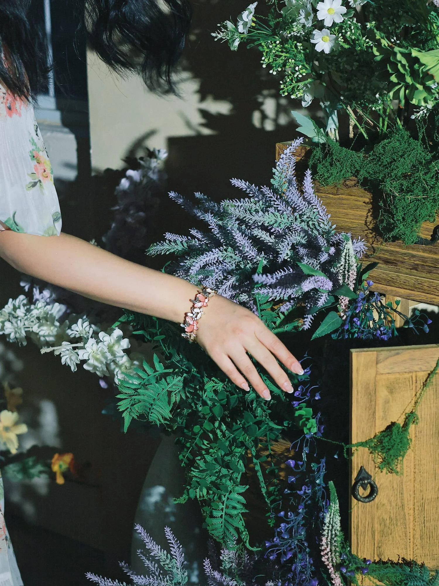 Wrist wearing the gold-toned enamel butterfly bracelet, hand reaching into lavender-coloured artificial flowers and ferns, white floral dress, dark hair visible.