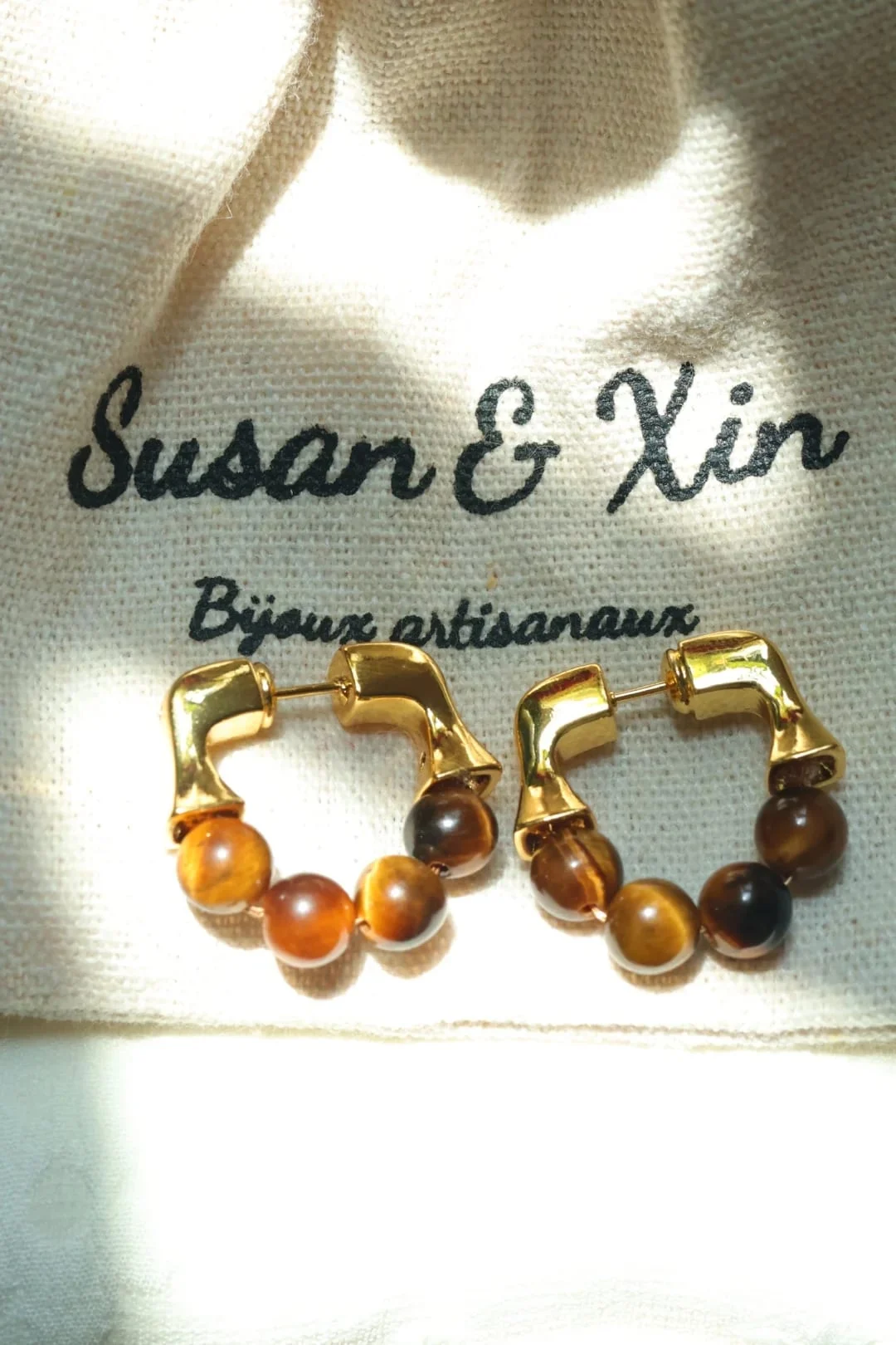 Pair of gold-toned hoop earrings with a squared upper section and four round tiger’s eye beads along the bottom of each hoop, on a Susan&Xin fabric pouch.