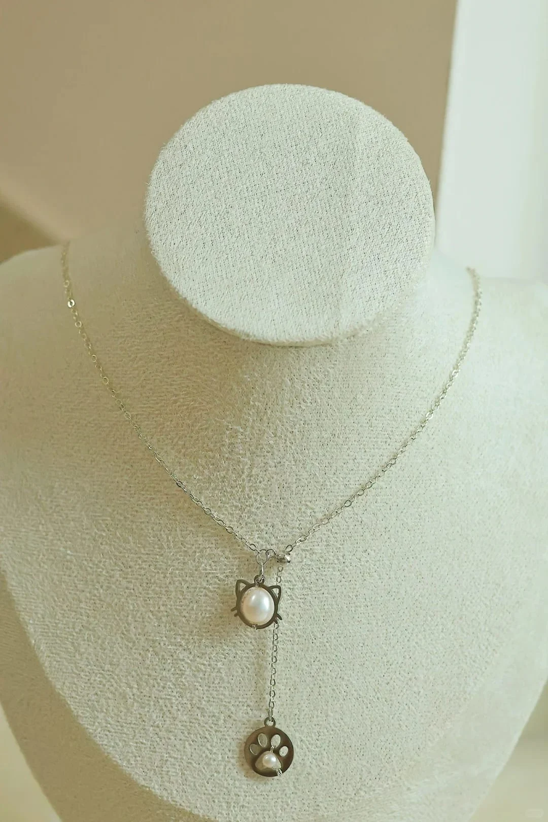 Fine silver-toned chain necklace on a display bust, with a cat-face frame holding a white iridescent oval bead and a chain drop ending in an openwork paw-print disc with a tiny white bead.