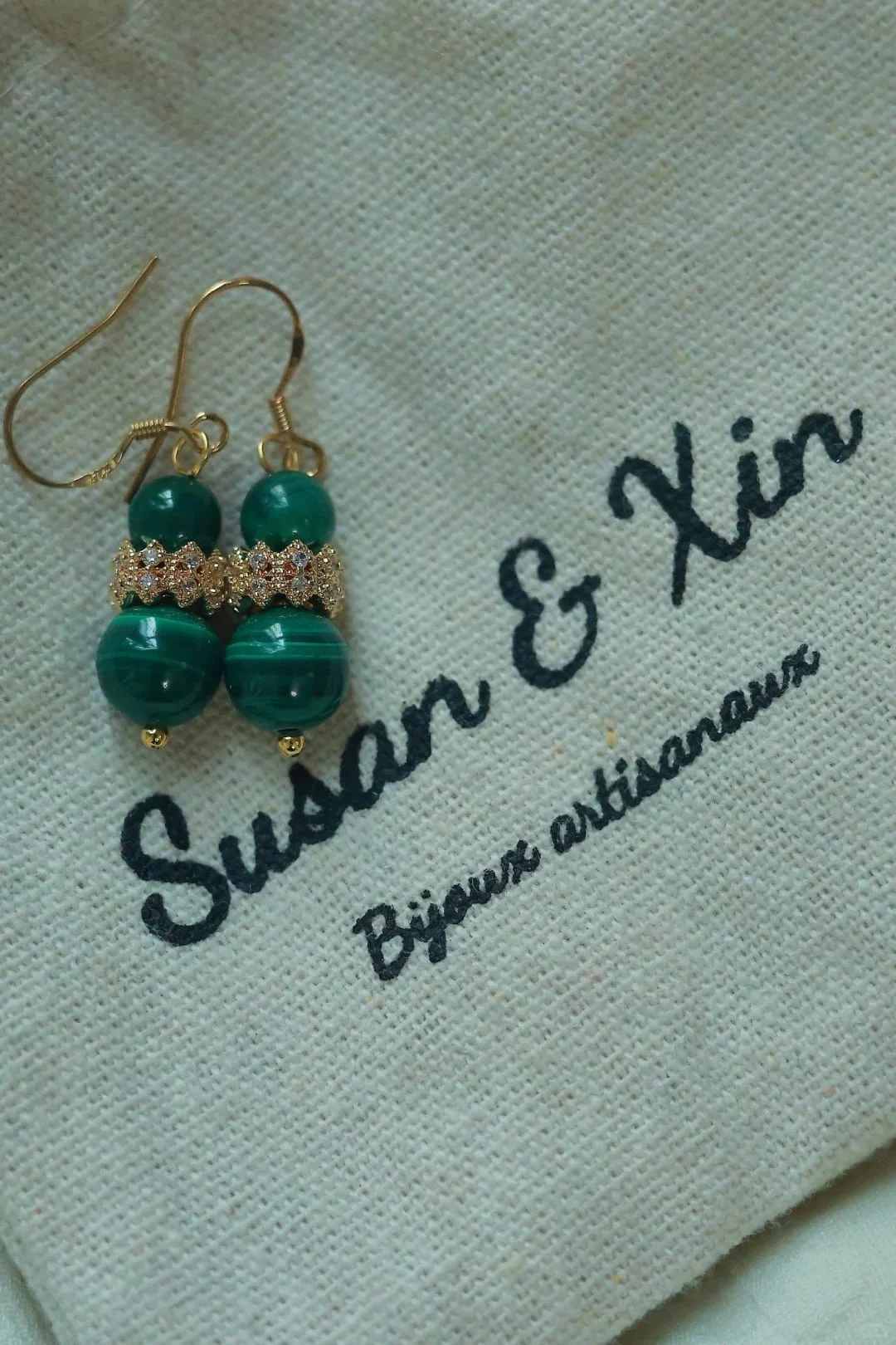 Pair of gold-toned hook earrings, each with a small malachite bead, a pavé-style gold-toned scalloped spacer and a larger malachite bead, on a Susan&Xin fabric pouch.