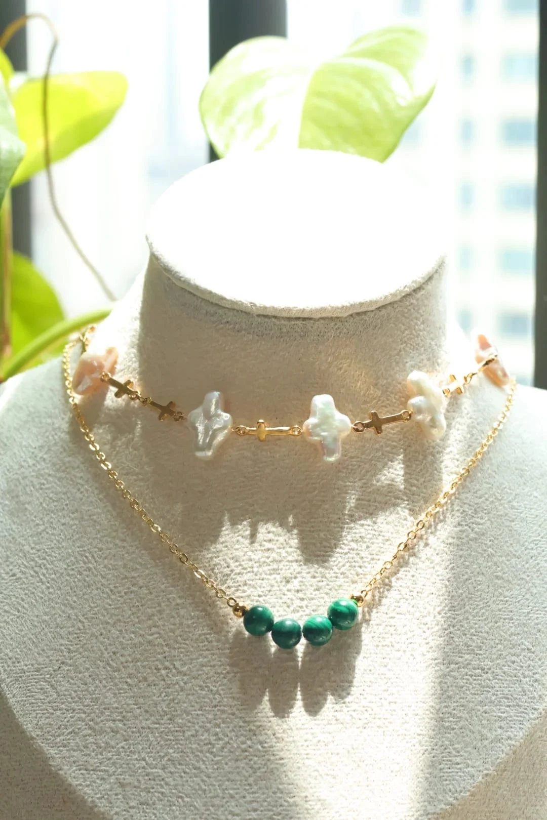 Two necklaces layered on a display bust by a window: a short chain of iridescent cross-shaped pieces and small gold-toned crosses above, and a fine gold-toned chain with four malachite beads below.