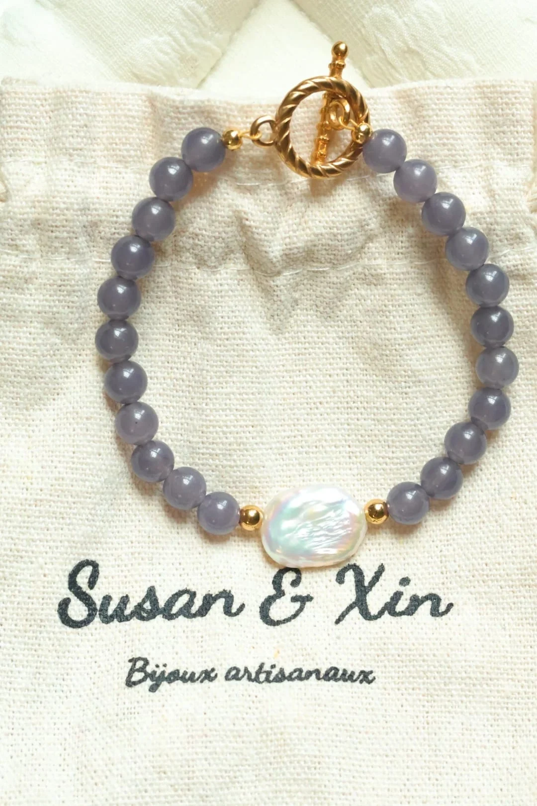 Grey-violet round-bead bracelet with a white iridescent centrepiece, small gold-toned beads and a toggle clasp on a Susan&Xin fabric pouch.