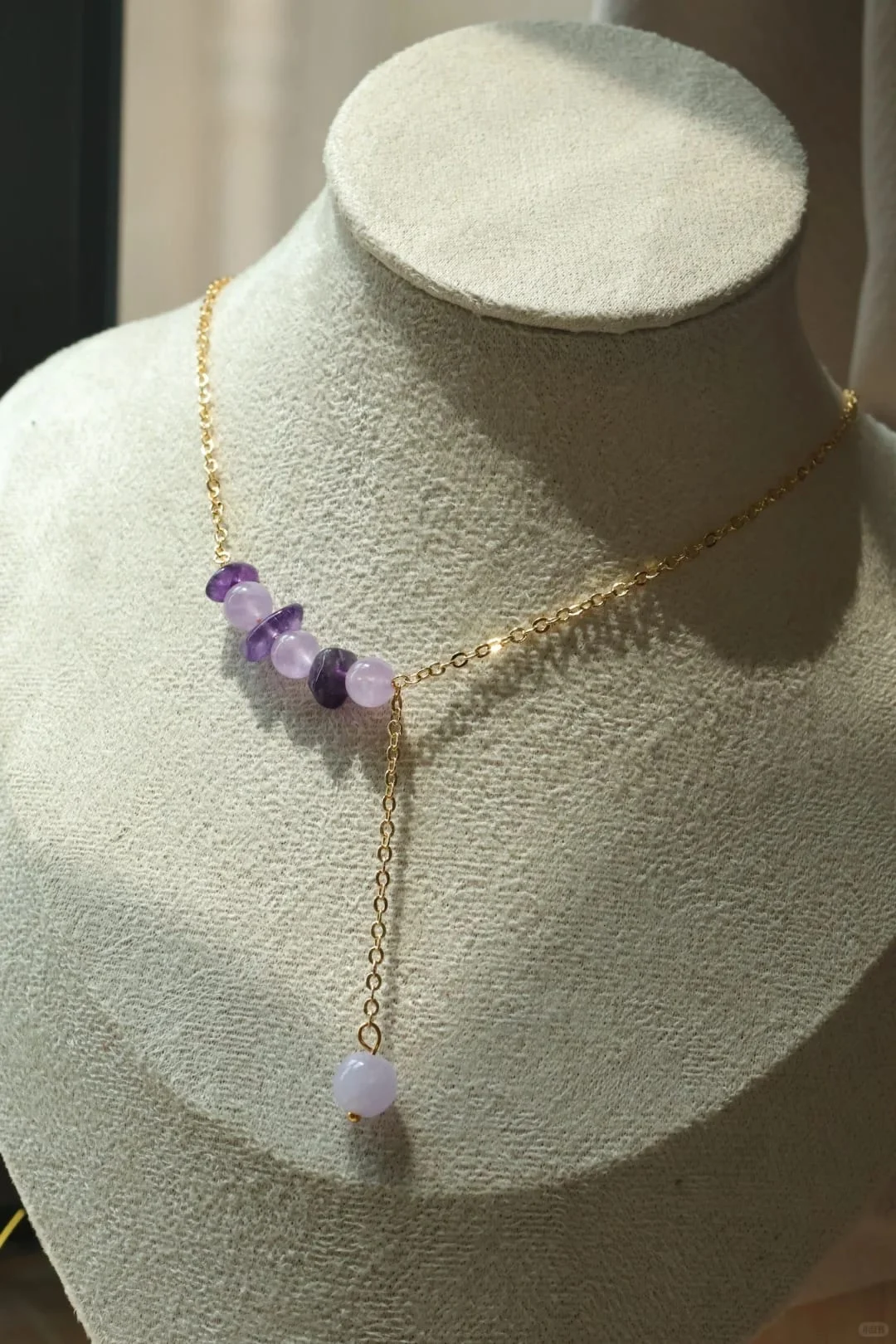 Gold-toned necklace on a display bust, with pale lilac and darker violet beads and a single-bead chain drop.