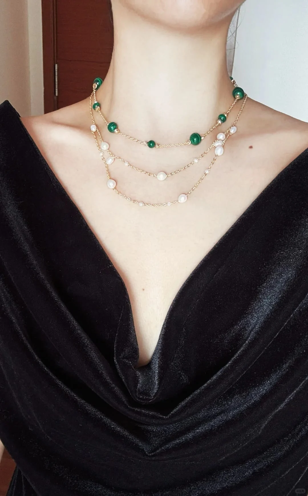 Model in a black velvet cowl-neck top wearing three layered necklaces: the malachite station necklace and two station necklaces with white iridescent beads.