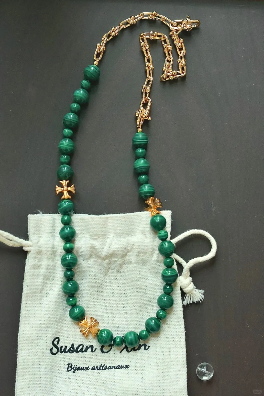 Necklace of banded malachite beads in alternating sizes with three gold-toned cross-shaped spacers and a length of chunky gold-toned U-link chain, on a Susan&Xin pouch against a dark surface.