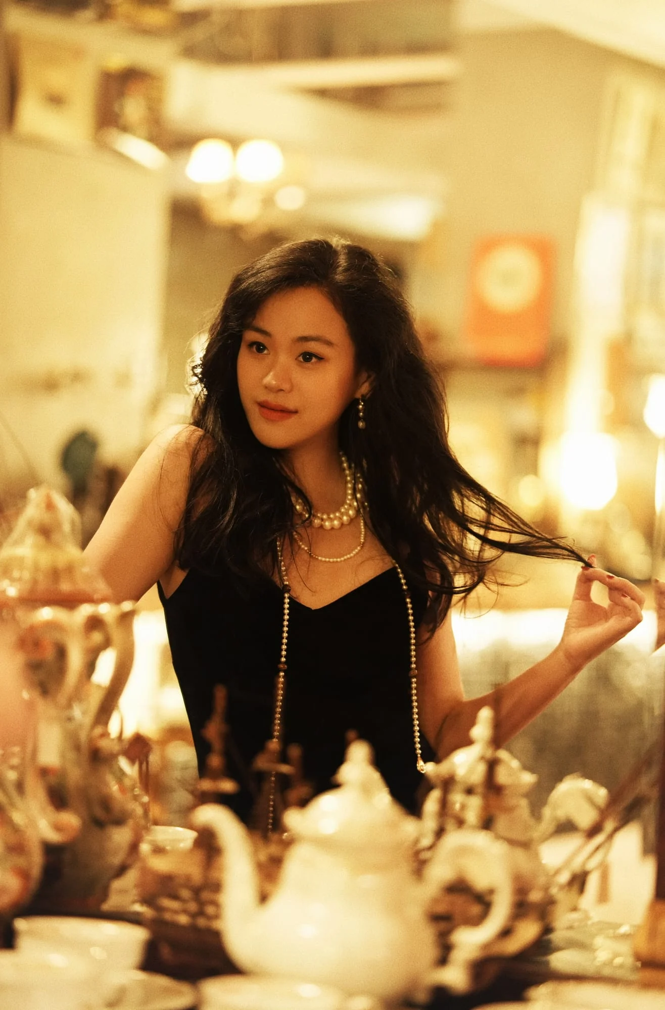 The long white-bead necklace with spaced brown oval beads hanging on a model in a black slip dress in a warmly lit tea room.
