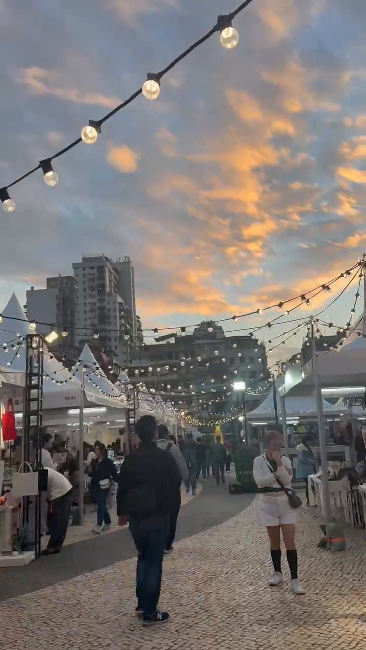 Market visitors beneath strings of lights and an orange evening sky in Macao.